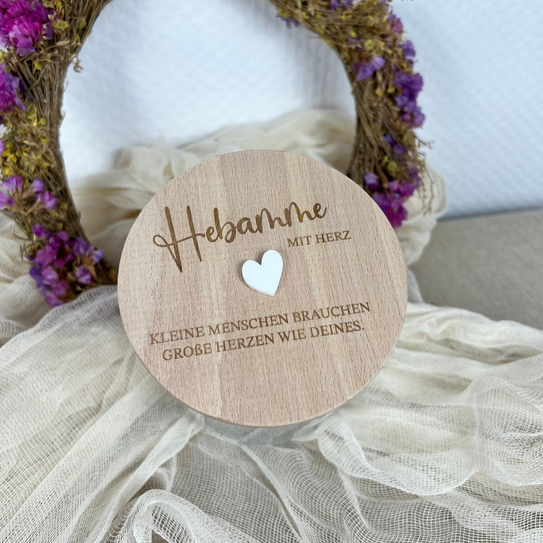 Glas mit Holzdeckel und Gravur „Hebamme mit Herz – Kleine Menschen brauchen große Herzen wie deines“, dekoriert mit Herzmotiv und Trockenblumen im Hintergrund