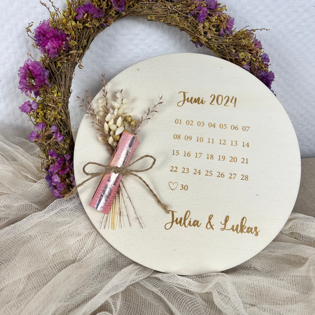 Runde Holzscheibe mit Gravur „Juni 2024“ und markiertem Hochzeitsdatum am 30. Juni, dazu die Namen „Julia & Lukas“ und ein Trockenblumenstrauß mit Geldschein – dekorativ arrangiert vor Trockenblumenkranz