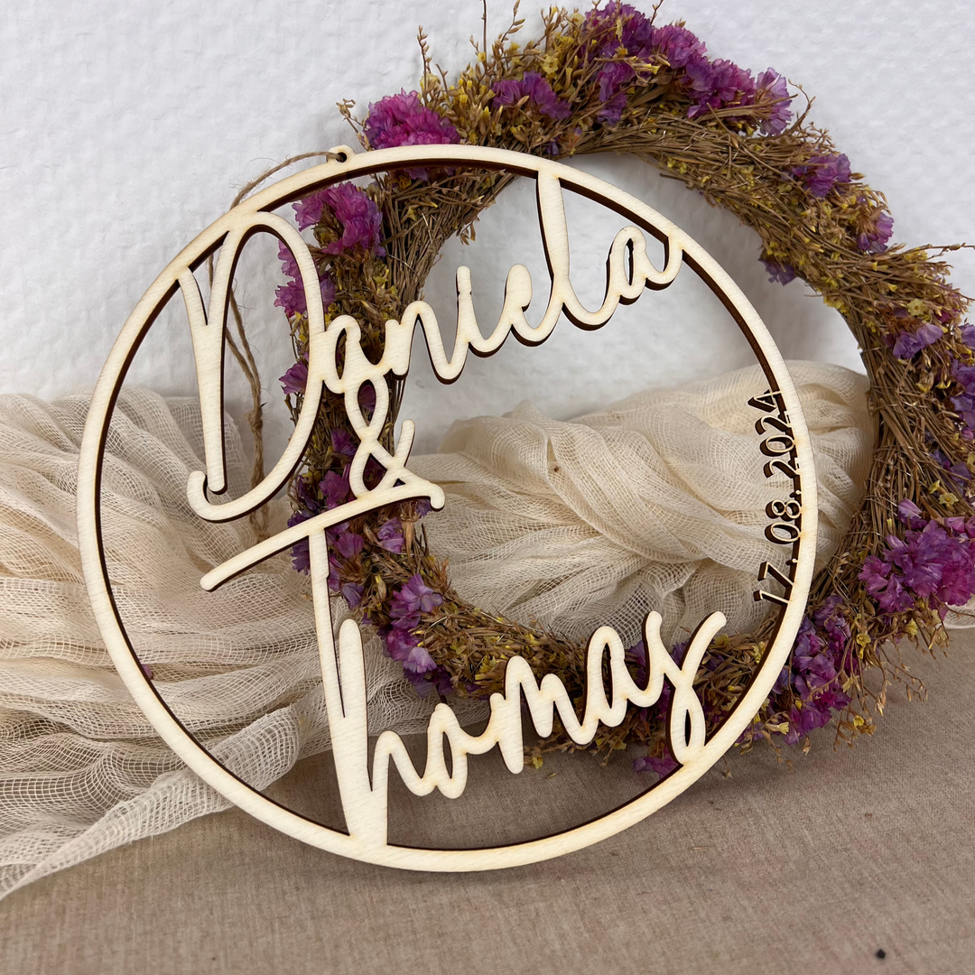 Rundes Holzschild mit den Namen „Daniela & Thomas“ und Hochzeitsdatum, ausgestanzt im Kreisdesign vor dekorativem Trockenblumenkranz